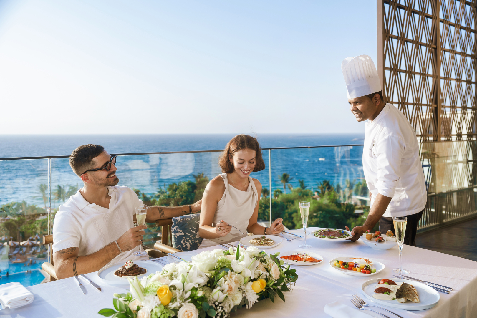 A couple enjoys a private fine-dining experience on a terrace overlooking the ocean, with a chef serving gourmet dishes and champagne, creating a romantic and elegant coastal ambiance.